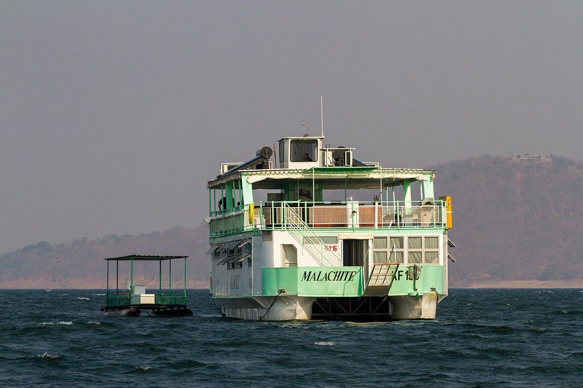 Malachite underway on Lake Kariba past dry escarpment hills and a tender boat.